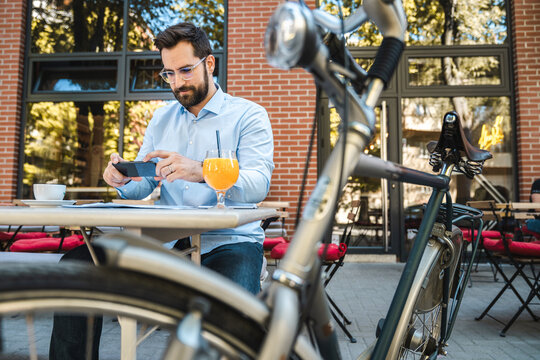 Businessman sitting and working in a local restaurant. He is sitting alone with a parked bicycle outside, drinking an orange juice and taking some photos with his phone.