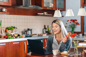 Young happy woman sitting at table while using laptop and eating breakfast in kitchen. Smiling...