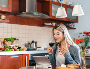 Young happy woman using tablet while drinking red wine in the kitchen. Satisfied blonde girl having a video call in her home during the dinner.