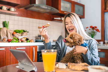 Young happy woman sitting at table and eating breakfast while holding a dog in her lap in kitchen. Hungry dog is trying to steal her food.