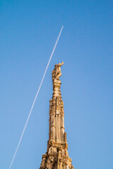 A spire and pinnacle sculptures of Milan Cathedral. Airplane leaving a line of cloud trail in the background.