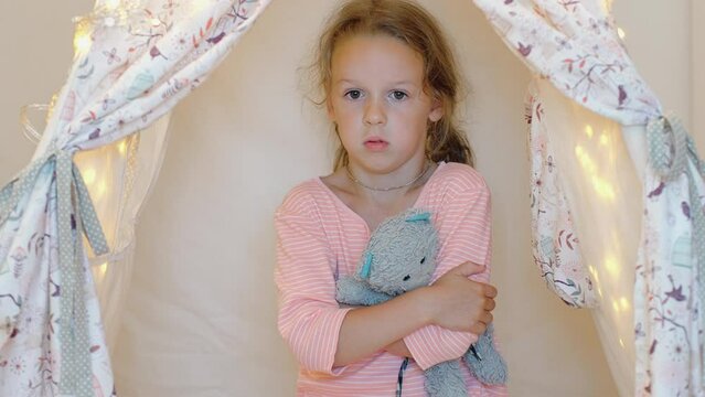 A frightened child hugs a teddy bear and listens to voices and sounds, a little girl alone in a cubby house, fear in the eyes of a defenseless child
