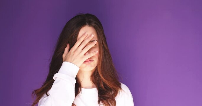 Young lady is shocked and disappointed making facepalm and rolling eyes in the studio on purple background. She is looking very upset, tired and angry. Woman wear white long sleeve rolling eyes.
