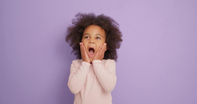Portrait of emotional African American excited amazed little girl kid looking shocked surprised at camera Girl with hands on cheek wide open mouth says wow isolated over purple background, Oh, my God
