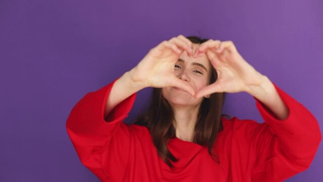 Happy attractive brunette woman look camera send blow air kiss showing shape heart with hands, girl wear red sweater isolated on purple background. Love, romantic concept.