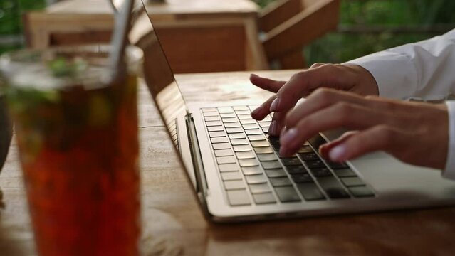 Woman typing on laptop keyboard in cafe. Writer creates a poem. Person composing email using notebook in outdoor coworking with ice tea, palm trees. Journalist writing article. Blogger making smm post