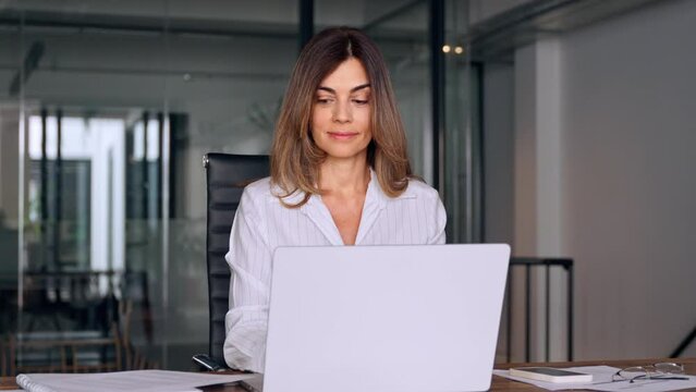 Smiling European Latin business woman accountant analyst holding documents, work at laptop computer doing online trade market tech research. Focused Hispanic businesswoman with paperwork in office.