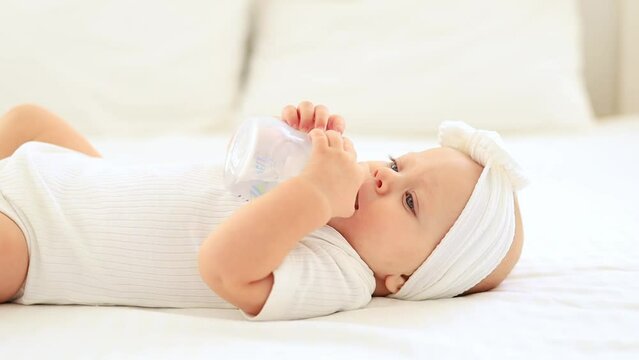 baby girl with blue eyes drinks milk or water from a bottle by herself holding it with her hands in a white bodysuit on a bed in a bright bedroom, baby food concept