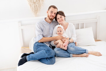Obraz premium a happy young family with a newborn baby hugging and kissing on the bed at home in a bright room, mom, dad and a small child on a white bed smiling
