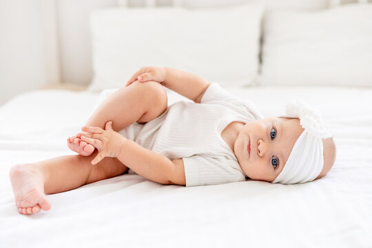 the baby is playing with his legs pushing them into his mouth, laughing happy little baby girl with blue eyes in a white bodysuit on a bed in a bright bedroom, a newborn baby is smiling happily