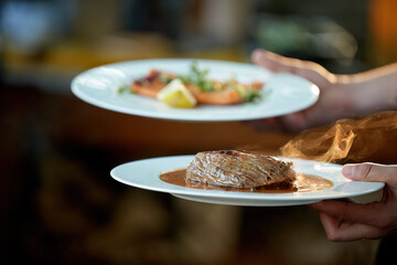 Serving food at the Garden Party.    Two plates in hand, with steak and salmon, in coloured, blurred backlight, shallow depth of field, backlit steam rising from the food.