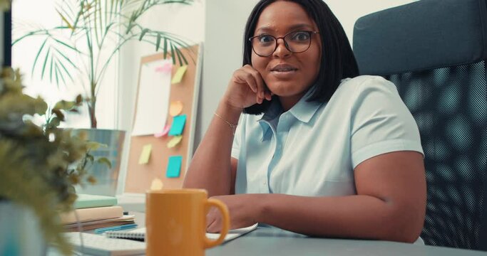 successful smiling black woman in white blouse sitting at table at workplace looking at camera, slow motion, favourite job, excited emotional manager loves, likes her profession