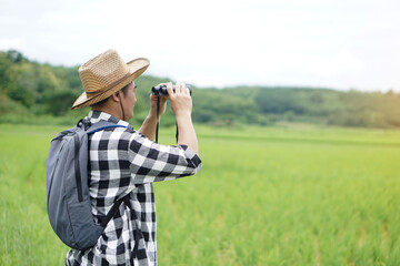 Asian man naturalist wears hat, plaid shirt, backpack, uses binocular to explore nature at paddy field. Concept, nature exploration. Ecology study. Pastime activity, lifestyle. Explore environment.   
