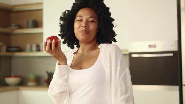 Portrait of beautiful young curly woman enjoying beautiful morning after shopping in supermarket and eating fresh red apple while looking at the camera at home kitchen Healthy food concept