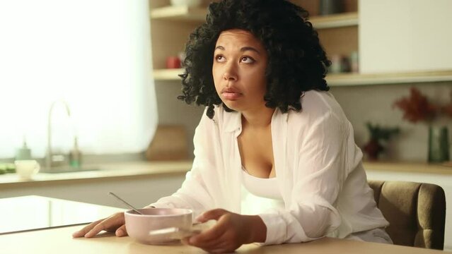 Boring exhausted curly woman watching uninteresting online videos programs or news in social media on smartphone gadget while having breakfast in morning at home Tired female after bad sleep insomnia