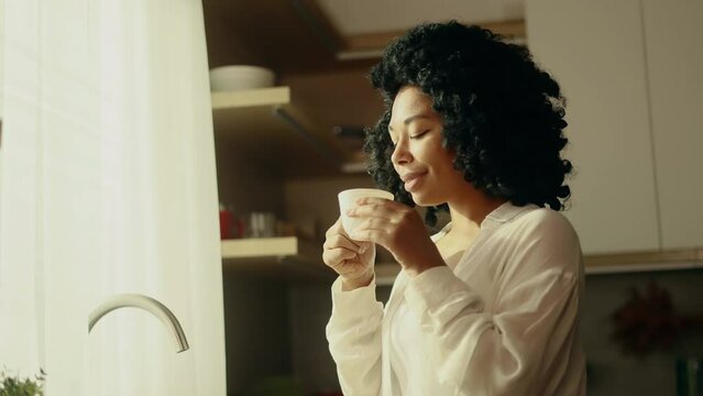Portrait of beautiful young curly woman looking out the window drinking hot drink of tea or coffee while the rays of the rising sun fall on her face at home kitchen Beautiful morning concept