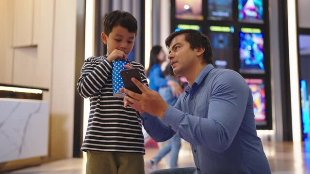 Asian father and little son using smartphone buying movie ticket at cinema theater. Father and son spending time on weekend togerther reserve seats in cinema via smartphone. Family activity concept