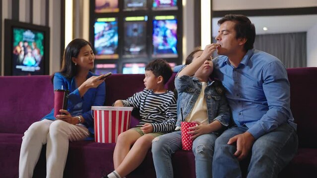 Happy Asian family enjoy eating popcorn and waiting for watching movie at front of cinema. Happy family waiting movie enjoy eating popcorn while sitting at sofa seat in front of cinema