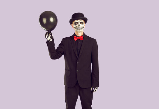 Black, mourning balloon in hands of artist playing zombie that has risen from the dead. Sad skeleton stage make-up on his face. Gloomy image for halloween on light purple background.