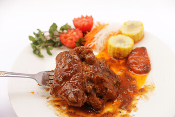A fork is placed beneath a pork rib stewed in a thick, aromatic gravy with vegetables and corn. Carrot, White Radish, Sliced Watercress tomatoes placed below. beside the tiled plate