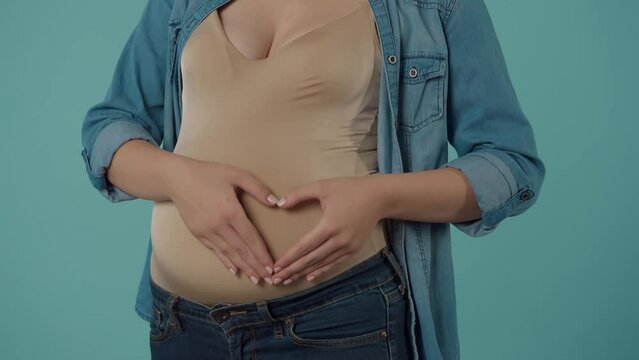 A pregnant woman strokes her bloated belly with love and tenderness, shows a heart sign, putting her hands on her stomach. Belly of a pregnant woman in the studio on a blue background close up.
