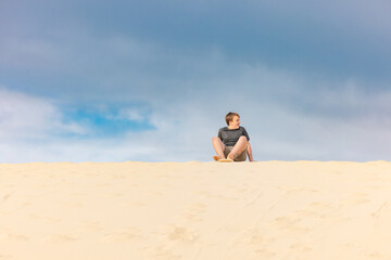 Boy sitting on sand board at the top of a dune ready to ride down with blue sky background