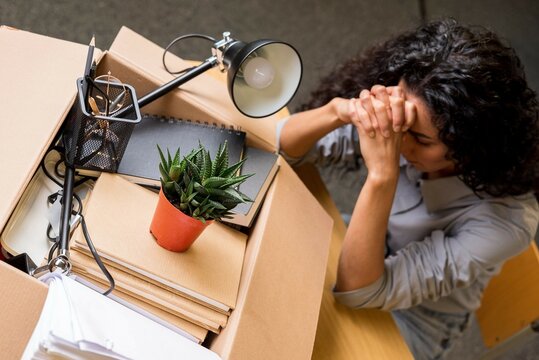 Photo top view. Curly-haired Spanish lady sits in stress leaning his head on his hands. There is a box with packed office supplies on the table. Unemployment and dismissal concept