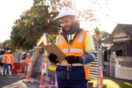 man road worker with beard and white hat wearing yellow jacket looking at his notes