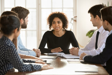 Multiracial businesspeople sit gather at office desk talk discuss business ideas at meeting together, diverse colleagues brainstorm at briefing, consider paperwork document, cooperation concept