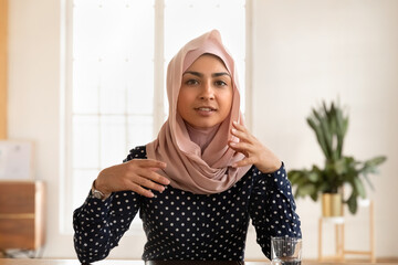 Portrait of young indian woman wearing hijab sit at desk talk on video call have interview conversation online, millennial Arabic ethnic female engaged in webcam virtual conference in office