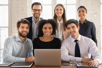 Group portrait of smiling multiracial young employees sit at desk in office cooperate on laptop, happy diverse colleagues coworkers work together on computer, collaboration, teamwork concept