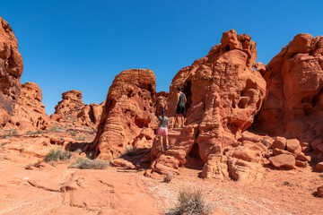 Fototapeta premium Couple at the entrance of windstone arch (fire cave) in Valley of Fire State Park, Mojave desert, Nevada, USA. Scenic view of beehive shaped red sandstone rock formations. Barren deserted landscape