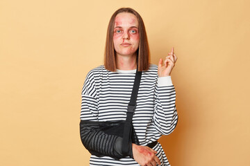 Serious sad woman in striped shirt with bandage and bruised skin standing isolated over beige background crossed finger trying to avoid violence having hopeful expression.
