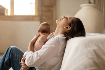 Tired young mother suffering from experiencing postnatal depression, sitting on floor and holding newborn baby on hands. Single mom, motherhood stressful