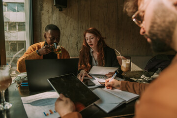 Good looking black male person is taking a sip of his tea while his ginger female colleague is explaining something to him. White man writing notes down in a notebook