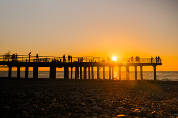 Silhouettes of people are walking on the pier against the clear sky over the Black Sea at sunset. Summer, peaceful, vacation and sightseeing concept