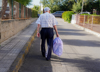 An old man walking on the street carrying a rubbish bag. Elderly recycle garbage