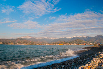 View of the blue sky with white clouds over the mountains in Batumi, Georgia. Waves of the Black Sea are crashing on the pebble beach - wide angle view