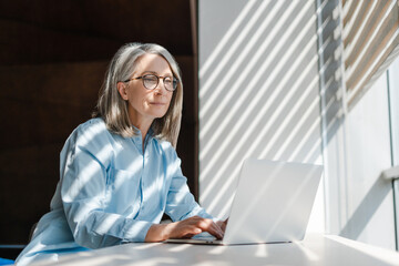Pensive senior businesswoman, successful gray haired manager using laptop computer working online...