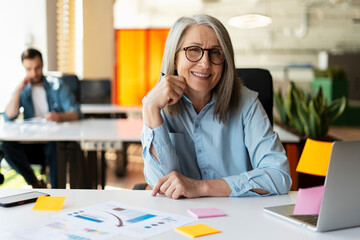 Portrait of smiling confident senior businesswoman using sticky notes looking at camera sitting in...
