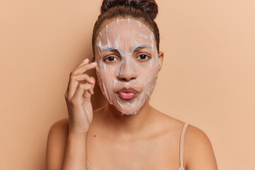 Beauty and cosmetology concept. Indoor close up of young pretty concentrated European lady standing isolated in centre on beige background applying face sheet mask to make skin fresh and smooth