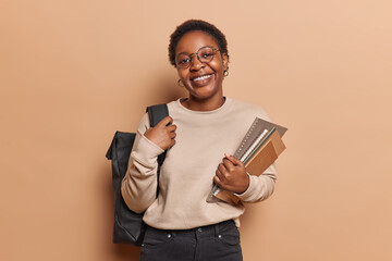 Cheerful dark skinned college student poses with notepads and rucksack on shoulder returns home...