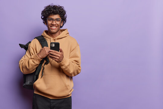 People and happiness. Studio photo of young cheerful smiling Hindu male student wearing beige hoodie and black trousers standing on left isolated on purple background holding smartphone with space
