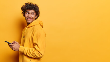 Cheerful Hindu man using cellphone and smiling reading good news message enjoying mobile application looks behind dressed in casual sweatshirt isolated over yellow background copy space for your promo
