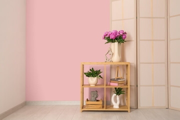 Vase of pink peonies and figurine on shelving unit with dressing screen near color wall