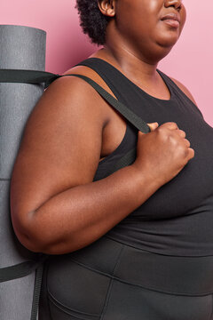Cropped shot of unrecognizable obese woman carries rubber rolled karemat on shoulder dressed in black sportswear ready for fitness training or workout poses against pink background. Healthy lifestyle