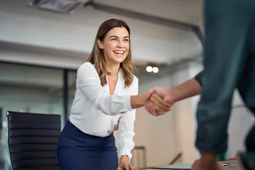 Foto auf Acrylglas Höhenskala Happy mid aged business woman manager handshaking greeting client in office. Smiling female executive making successful deal with partner shaking hand at work standing at meeting table.