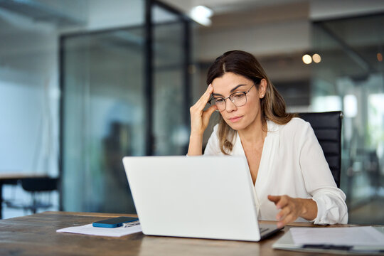 Worried fatigued mature business woman wearing glasses having headache at work. Tired busy 40s middle aged businesswoman feeling stress at workplace looking at laptop computer in office.