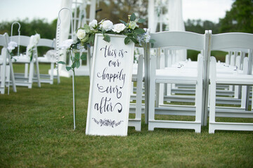 White sign that reads "this is our happily ever after"at top of wedding ceremony isle with white chairs and white alter