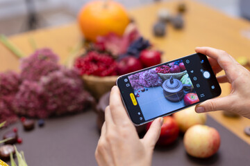 A girl shoots a background with an autumn harvest on her phone. The blogger takes photos and videos on the phone. Hands with phone close-up. Autumn vegetables and fruits on a brown background.